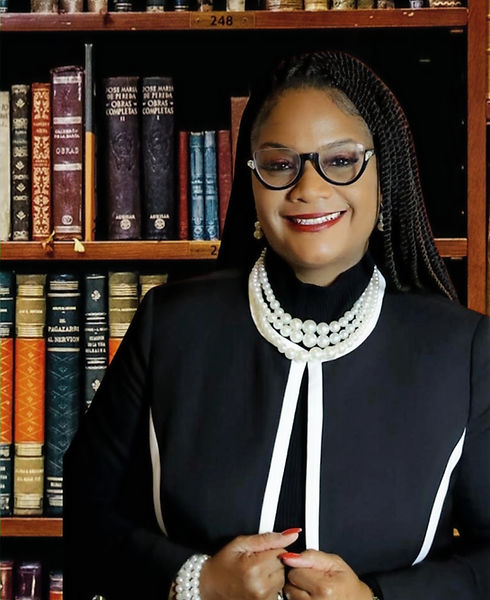A woman wearing glasses, a black and white outfit, and pearl jewelry stands smiling in front of a bookshelf filled with old books.
